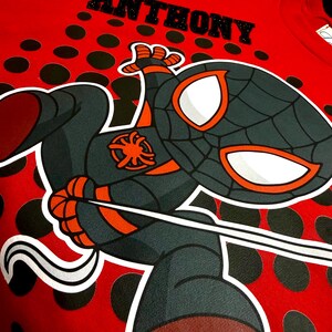 May include: Red t-shirt featuring a cartoon Spider-Man character with the name "ANTHONY" above. The character is in black, gray, and orange, with white eye details. The shirt has a polka dot pattern.