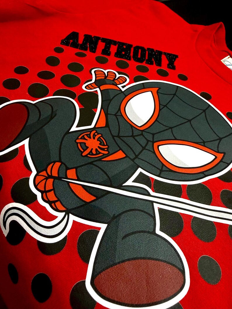May include: Red t-shirt featuring a cartoon Spider-Man character with the name "ANTHONY" above. The character is in black, gray, and orange, with white eye details. The shirt has a polka dot pattern.