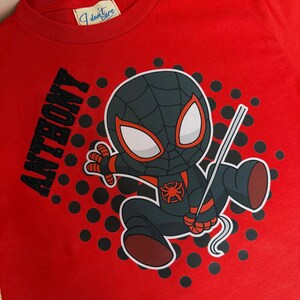 May include: Red t-shirt featuring a cartoon Spider-Man character in a black and gray suit with red accents. The name "ANTHONY" is printed in black on the left side. The shirt has a round neck and a tag that says "I don't care".