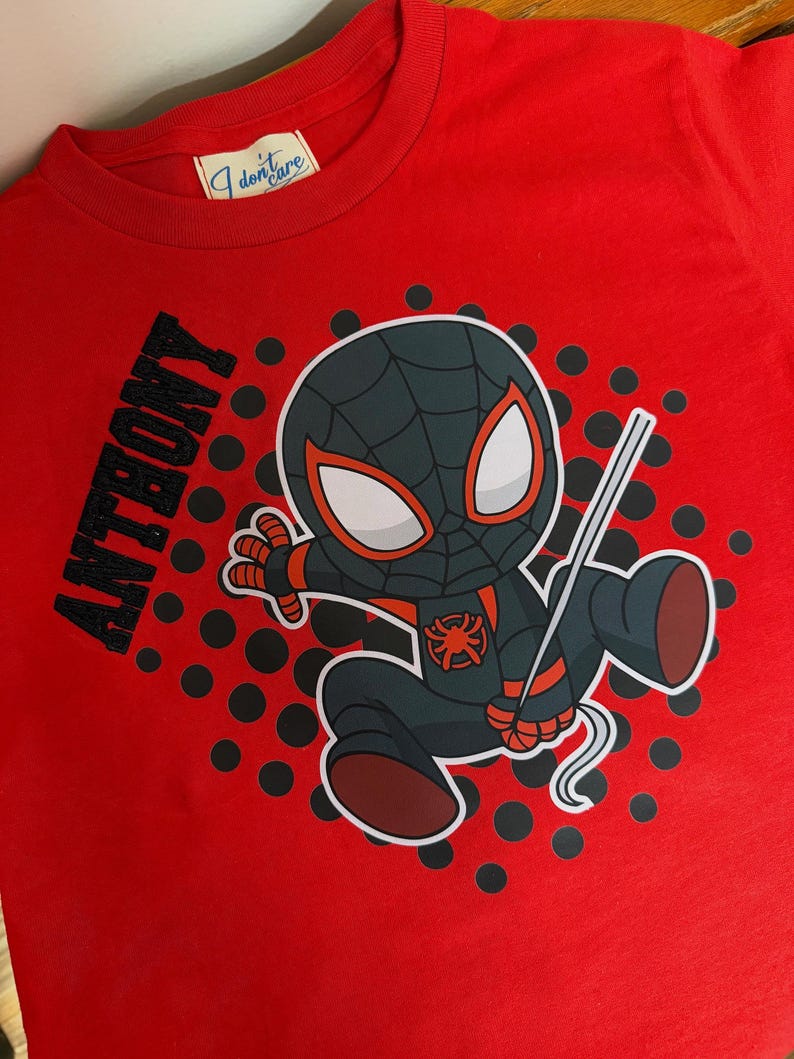 May include: Red t-shirt featuring a cartoon Spider-Man character in a black and gray suit with red accents. The name "ANTHONY" is printed in black on the left side. The shirt has a round neck and a tag that says "I don't care".