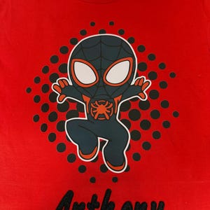 May include: Red t-shirt featuring a cartoon Spider-Man character in a black and blue suit with orange accents. The shirt has the name "Anthony" embroidered in black script at the bottom. A pattern of black dots surrounds the character.