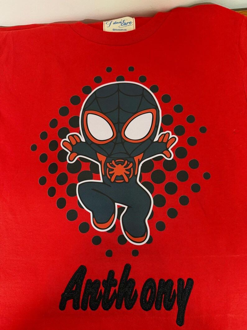 May include: Red t-shirt featuring a cartoon Spider-Man character in a black and blue suit with orange accents. The shirt has the name "Anthony" embroidered in black script at the bottom. A pattern of black dots surrounds the character.