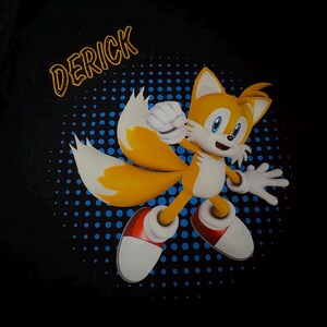 May include: Black t-shirt featuring a cartoon character with orange fur, two tails, and white gloves and shoes. The character is set against a blue dotted background. The name "DERICK" is printed in gold letters above the character.