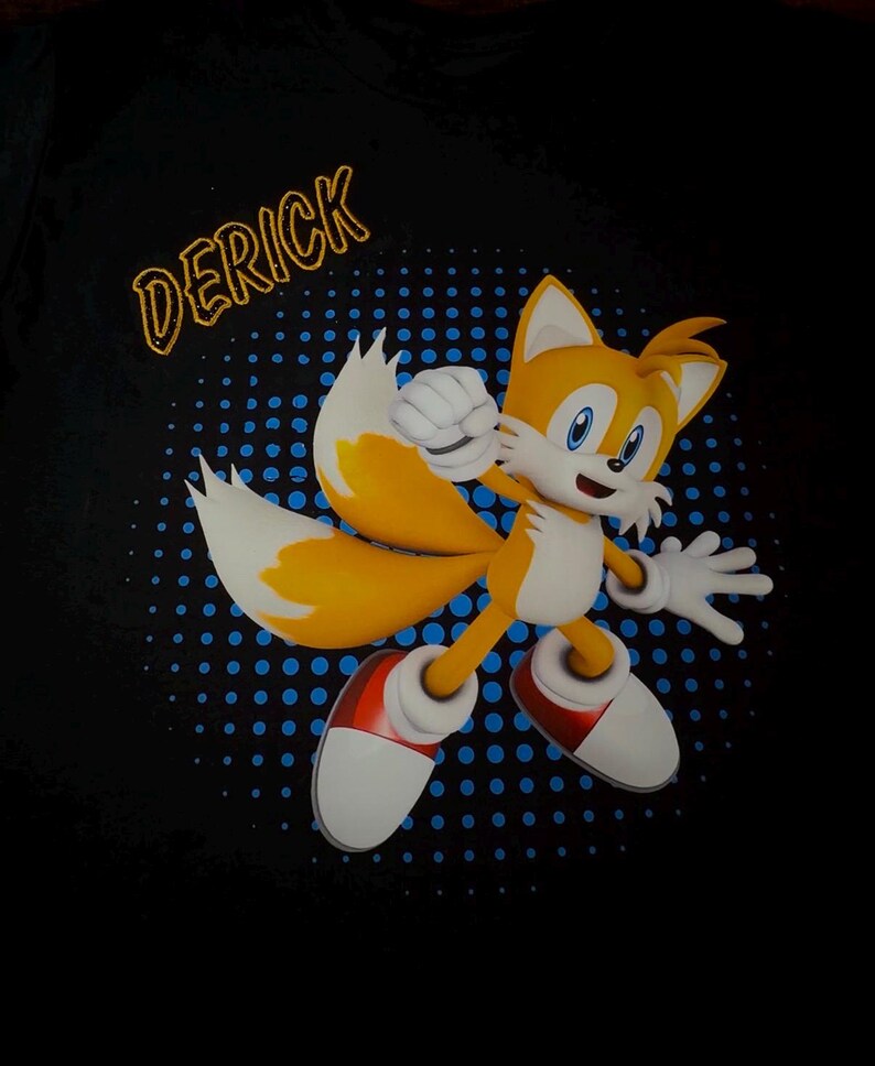 May include: Black t-shirt featuring a cartoon character with orange fur, two tails, and white gloves and shoes. The character is set against a blue dotted background. The name "DERICK" is printed in gold letters above the character.