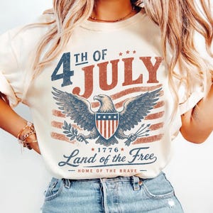 Puede incluir: Una camiseta de color crema con un gráfico desgastado que muestra un águila calva, un escudo y el texto 4TH OF JULY, 1776, LAND OF THE FREE y HOME OF THE BRAVE en rojo, blanco y azul, con un diseño de bandera estadounidense descolorida.