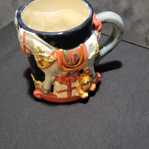 May include: A ceramic mug with a rocking horse design. The mug features a white horse with a red saddle, a blue handle, and a teddy bear. The mug is decorated with Christmas-themed elements.