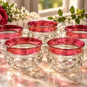 May include: A set of five clear glass cups featuring a textured, circular pattern on the exterior and a wide, vibrant magenta-coloured rim. Each cup includes a small, curved handle attached to the side, showcasing a consistent design across all pieces.