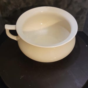 May include: Cream-colored ceramic chamber pot with a handle. The pot has a rounded body and a wide, flat rim. The handle is small and curved. The chamber pot is sitting on a dark surface.