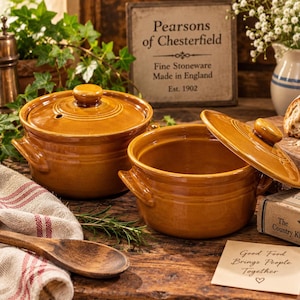 May include: Two round, glazed mustard-yellow stoneware pots with matching lids and side handles. Each pot features a circular knob on the lid and a smooth, glossy finish. The pots are arranged together, with one lid placed beside its open pot.