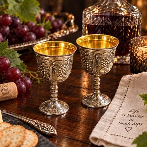 May include: Two silver-toned metal goblets with gold-toned interiors, featuring embossed grape vine patterns on the exterior. The goblets stand on a dark wooden surface alongside a cork printed with Good Wine Good Company and a napkin with the text Life is Sweeter in Small Sips.