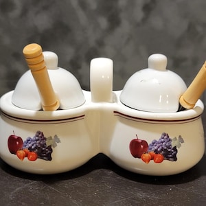 May include: White ceramic double condiment server with lids and wooden spoons. The server features a handle and is decorated with a fruit design of apples and grapes. A decorative kitchen item.