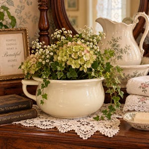 May include: A cream-colored ceramic chamber pot filled with green hydrangea blossoms, white baby's breath, and trailing ivy vines. The pot features a single side handle and sits on a white lace doily next to stacked books and a floral pitcher.