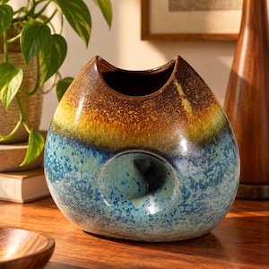 May include: A rounded ceramic vase featuring a smooth, glossy finish with a color gradient transitioning from a deep brown at the top to a mottled blue at the base. The vase has a distinct circular indentation on the front and a curved rim.