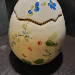 May include: A ceramic egg-shaped container with a lid, hand-painted with delicate floral designs. The flowers are in shades of white, pink, and blue, with green leaves. The container is on a dark surface.