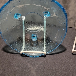 May include: A clear, blue glass plate with a unique design. The plate has a circular shape with a raised rim and three decorative glass elements. The plate is supported by a white metal stand.