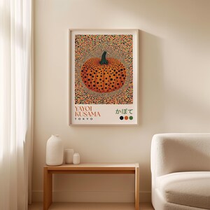 Yayoi Kusama–style pumpkin poster displayed above a wooden bench with white ceramic vases, beside a soft neutral sofa. The central design features an orange pumpkin with dark black dots and a green stem, set within a dotted pattern that radiates outward in red, orange, and green. The white section at the bottom contains “YAYOI KUSAMA TOKYO,” Japanese characters, and color dots in coordinating tones, balancing modern and organic aesthetics.