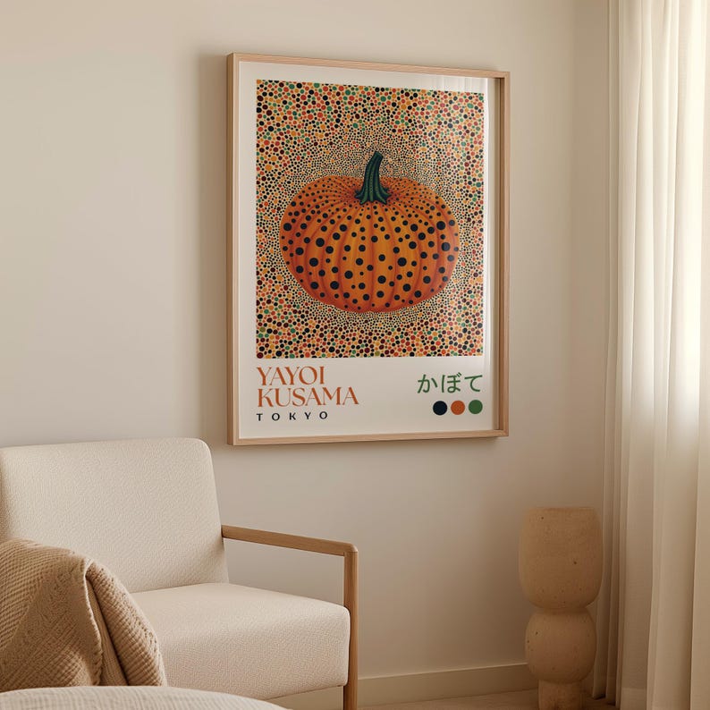 Yayoi Kusama–style pumpkin poster hanging on a neutral wall beside a cream armchair and sheer curtains. The artwork shows a round orange pumpkin with black polka dots and a green stem, surrounded by a vibrant dotted background in orange, green, and red tones. The lower white border includes “YAYOI KUSAMA TOKYO,” Japanese text, and three matching color swatches, giving the piece a clean, graphic finish.