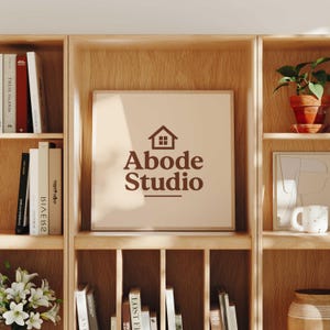 Abode Studio logo displayed on a framed print with a brown house icon and serif lettering, styled on a wooden bookshelf surrounded by books, plants, and home decor in a warm, cozy interior