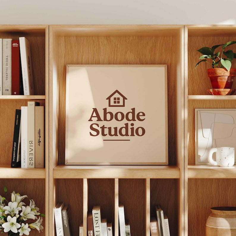 Abode Studio logo displayed on a framed print with a brown house icon and serif lettering, styled on a wooden bookshelf surrounded by books, plants, and home decor in a warm, cozy interior