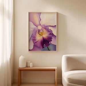 Large floral artwork of a white and purple orchid with a golden yellow center, framed in light wood and leaning against a white wall on a wooden floor. Soft natural light casts shadows across the floor and wall.