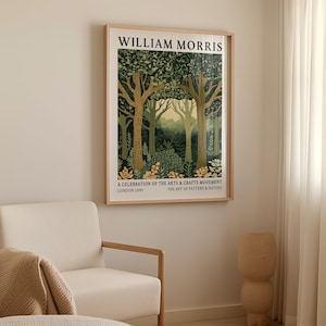 William Morris forest scene art print in a natural wood frame displayed above a light beige armchair. The artwork depicts a trio of elegant trees, their branches stretching upward and crowned with a canopy of patterned leaves in deep and muted greens. Soft golden accents highlight the forest floor, while a distant pale yellow sky glows between the trees. The design is framed with bold typography at the top and historical reference text at the bottom.