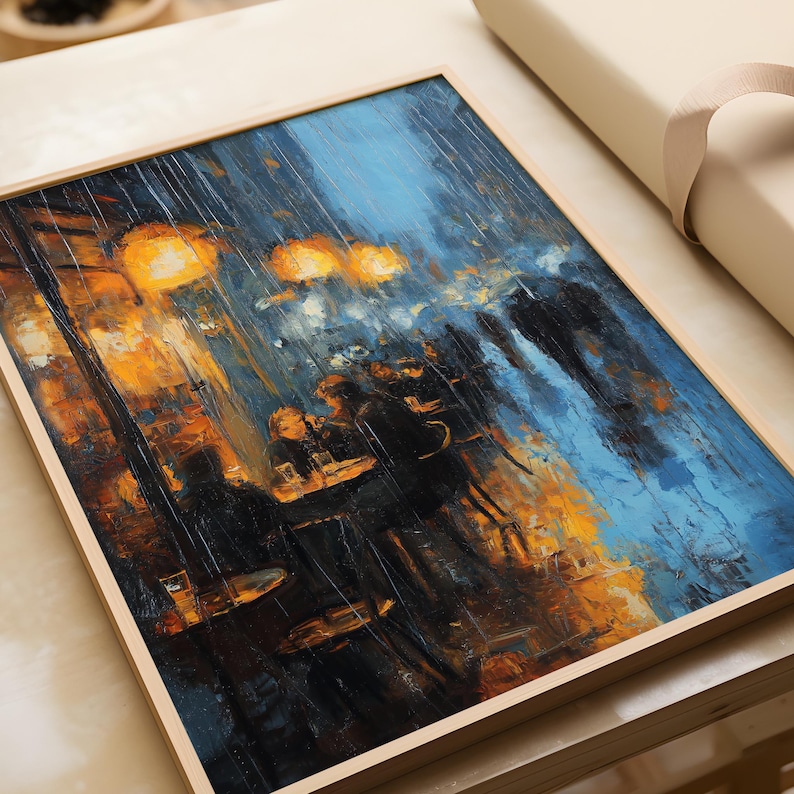 Close-up of the Monet poster laid flat on a table, partially wrapped in kraft paper for packaging. The artwork depicts glowing café lights in rich golden tones, contrasting with deep navy and cerulean blues of the rainy street. Figures at tables and passing silhouettes are softened by heavy brushstrokes and vertical rain textures. The natural wood frame adds warmth to the atmospheric impressionist print.