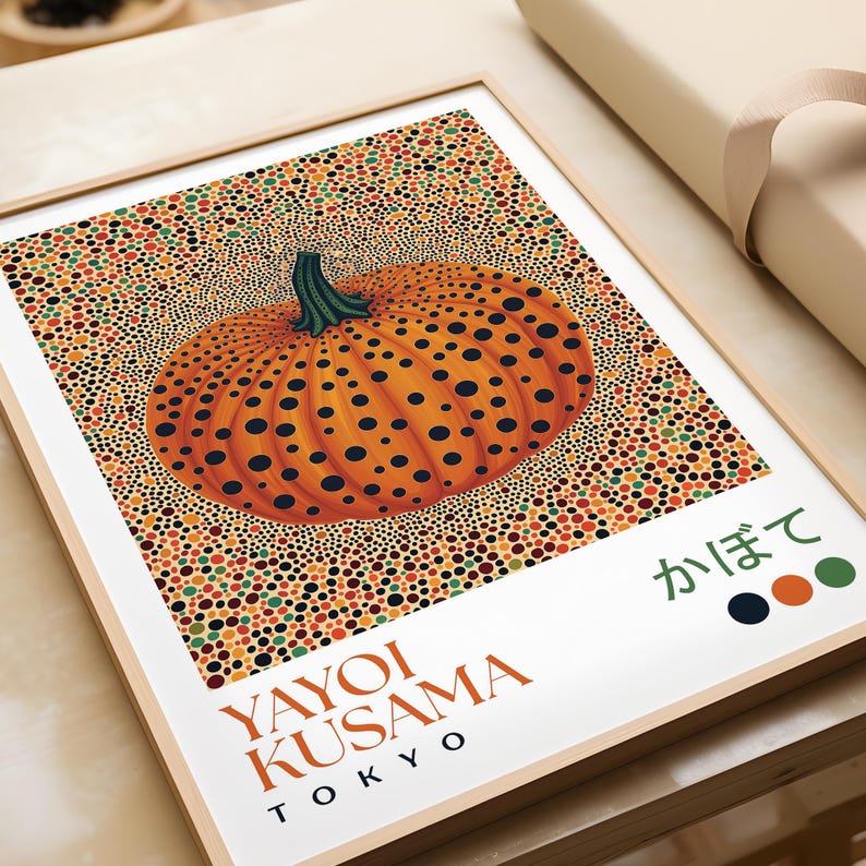 Close-up of a Yayoi Kusama–inspired pumpkin print lying flat on a tabletop. The composition centers on an orange pumpkin with deep black dots and a textured green stem, surrounded by a dense field of multicolored dots in orange, red, yellow, and green. The lower white edge features “YAYOI KUSAMA TOKYO,” Japanese text, and color swatches, highlighting the intricate details and bold contemporary pop-art style.