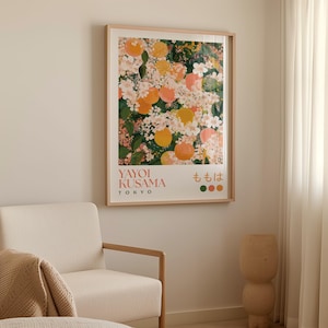 Yayoi Kusama-style poster of peach blossoms and fruit in a warm peach and green color palette, hanging above a light armchair and sculptural beige side table in a softly lit interior with sheer curtains.