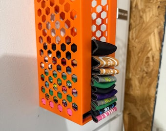 May include: An orange, honeycomb-patterned organizer attached to a white refrigerator. The organizer holds folded socks of various colors and patterns. The design features a slotted front and a side compartment for storage.