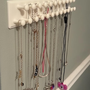 May include: A white wall-mounted jewelry organizer with multiple pegs for hanging necklaces. Various necklaces of different lengths and styles, including silver, gold, and black chains, are displayed. The organizer is mounted on a light gray wall.