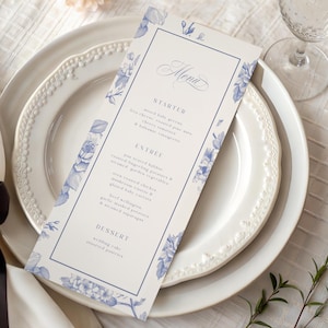 Op de afbeelding: Een trouwmenu met een blauw bloemmotief en het woord "Menu" in elegant schrift. De kaart ligt op een wit bord met een decoratieve rand, met een wijnglas op de achtergrond. Het menu bevat voorgerechten, hoofdgerechten en desserts.