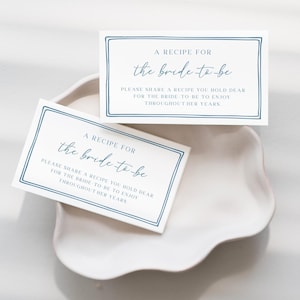 Op de afbeelding: Twee witte receptkaarten met blauwe tekst en randen worden weergegeven op een wit, geschulpte schaal. De kaarten luiden "A Recipe For the bride to be" en nodigen gasten uit om recepten voor de bruid te delen.
