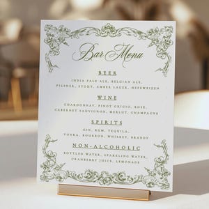 Könnte beinhalten: Eine weiße Barkarte mit grünen floralen Designs. Die Karte listet Bier, Wein, Spirituosen und alkoholfreie Getränke auf. Die Karte wird in einem goldfarbenen Metallständer präsentiert.