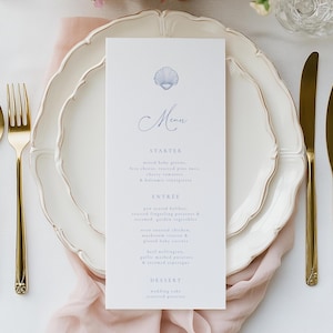 May include: A refined table setting featuring a white plate with a decorative edge, gold cutlery, and a menu card. The menu card displays a seashell illustration and the word "Menu". The menu lists starter, main course, and dessert options.