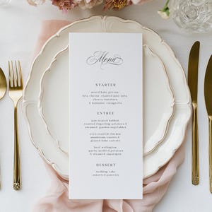 May include: Elegant wedding menu card with a floral design, placed on a white plate with a gold rim. The menu lists starter, entree, and dessert options. Gold silverware and a pink cloth complete the table setting.
