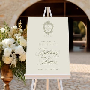 Könnte beinhalten: Ein cremefarbenes Hochzeits-Willkommensschild auf einer weißen Staffelei. Das Schild zeigt ein Wappen mit den Initialen "B" und "T", das Datum "09.10.28" und die Namen "Bethany" und "Thomas" in eleganter Schrift. Der Ort ist Oakdale, New York.
