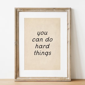Peut inclure: Impression encadrée avec l'inscription "you can do hard things" en noir, dans une police manuscrite. L'impression a un fond beige et est encadrée dans un cadre en bois marron clair vieilli. L'œuvre est exposée sur une surface en bois.