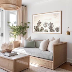 May include: A living room scene featuring a light-colored woven sofa with several pillows in shades of white and green. A framed black and white photograph of palm trees hangs above the sofa. A woven coffee table and a woven light fixture are also visible.