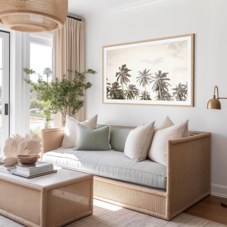 May include: A living room scene featuring a light-colored woven sofa with several pillows in shades of white and green. A framed black and white photograph of palm trees hangs above the sofa. A woven coffee table and a woven light fixture are also visible.