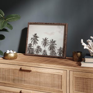 May include: A framed black and white print of palm trees. The print is in a brown wooden frame and sits on a light brown woven surface. A gold bowl with white and gold spheres, a small gold candle holder, and a white coral decoration are also on the surface.