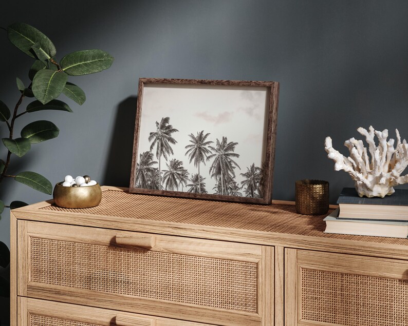 May include: A framed black and white print of palm trees. The print is in a brown wooden frame and sits on a light brown woven surface. A gold bowl with white and gold spheres, a small gold candle holder, and a white coral decoration are also on the surface.