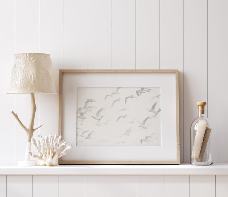 May include: A white wooden shelf with a white lamp with a cream-colored shade, a framed print of a flock of birds in flight, and a clear glass bottle with a cork stopper.