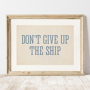 Puede incluir: Una impresión enmarcada con el texto "DON'T GIVE UP THE SHIP" en letras azules. La impresión tiene un fondo beige y está enmarcada en un marco de madera dorada. La obra de arte se exhibe contra una pared blanca.