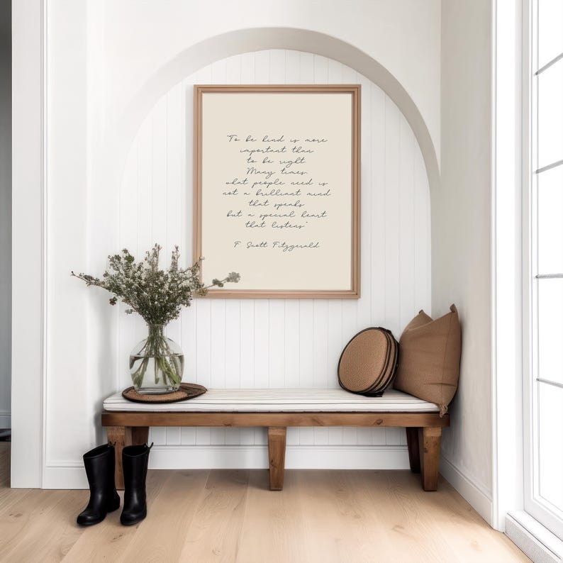 May include: A white wall with a framed quote by F. Scott Fitzgerald that reads "To be kind is more important than to be right. Many times people need not a brilliant mind that speaks but a special heart that listens." The quote is displayed above a wooden bench with a striped cushion and two brown throw pillows.