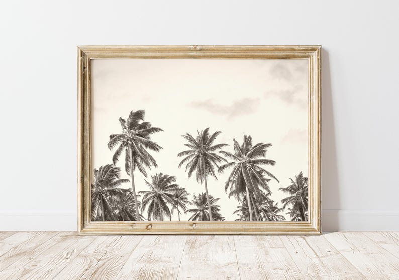 May include: Black and white framed print featuring a row of palm trees under a cloudy sky. The artwork is displayed in a light-colored wooden frame, adding a touch of warmth to the tropical scene. The print is set against a white wall.