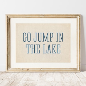 May include: A framed print with the text "GO JUMP IN THE LAKE" in blue letters. The print has a beige background and is encased in a gold-colored wooden frame. The artwork is displayed against a white wall.