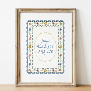 Könnte beinhalten: Gerahmter Kunstdruck mit dem Text "HOW BLESSED ARE WE" in einem dekorativen Oval. Der Druck hat einen blauen, wellenförmigen Rand mit floralen Akzenten in Rosa, Gelb und Grün. Der Rahmen ist hellbraun.