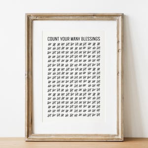 May include: Framed print with the text "COUNT YOUR MANY BLESSINGS" in black. The print features a grid of tally marks, also in black, on a white background. The frame is a light brown color.