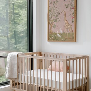 May include: A light wood baby crib with a white mattress and a light pink pillow. A beige blanket is draped over the side. A framed artwork with a giraffe and birds hangs on the wall above the crib.