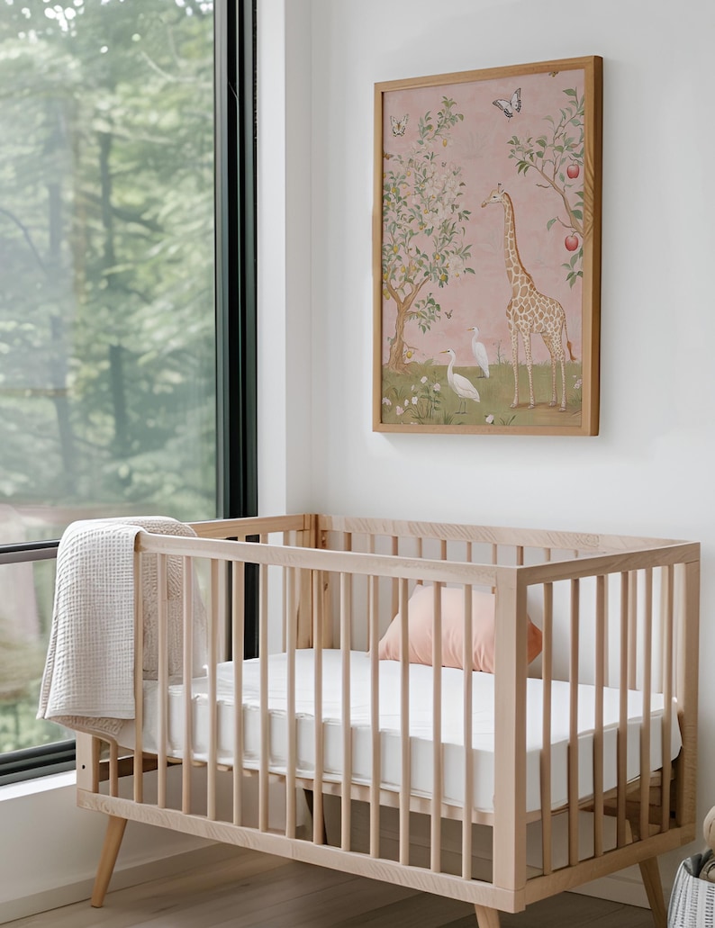May include: A light wood baby crib with a white mattress and a light pink pillow. A beige blanket is draped over the side. A framed artwork with a giraffe and birds hangs on the wall above the crib.